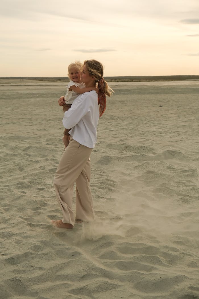 A mother and baby walking on a sandy beach during sunset, sharing a joyful moment.